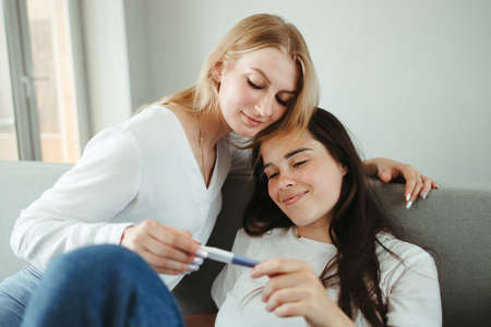 Two girlfriends holding pregnancy test sitting in living room. Homosexual family, lgbt equality, women supportの写真素材