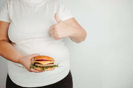 Balanced ration, diet, counting calories. Fast food and unhealthy snacks. Low calorie meals. Overweight woman with burger and thumb upの写真素材