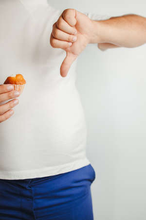 Compulsive overeating, weight loss, diet, unhealthy eating. Overweight man with cupcake and thumb downの写真素材