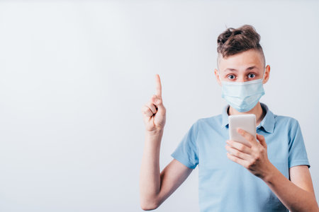 Insight. Important information. Attention gesture. Surprised boy in medical mask and gloves holding smartphone pointing finger up. Empty space for advertising.の写真素材