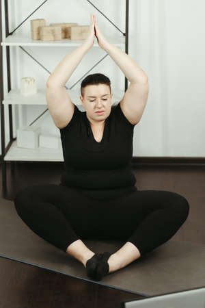 fitness, online personal trainer, home training, technology and diet. overweight woman doing yoga sitting on a fitness mat at home, using laptopの写真素材