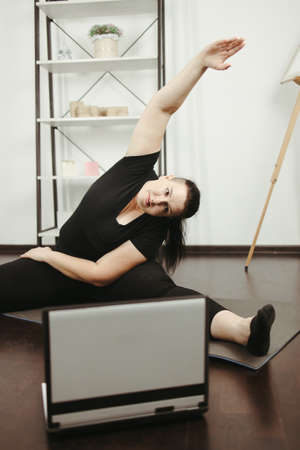 fitness, online personal trainer, home training, technology and diet. overweight woman doing stretching exercises sitting on a fitness mat at home, using laptopの写真素材
