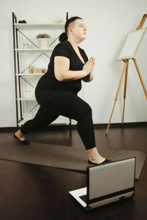 fitness, online personal trainer, home training, technology and diet. overweight woman doing yoga on a fitness mat at home, using laptopの写真素材