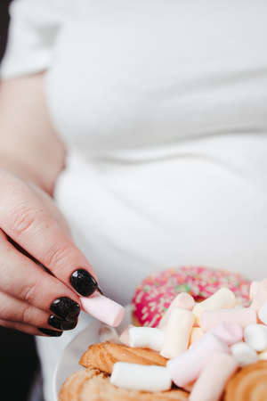 Weight loss, diet, unhealthy eating, overeating. Overweight woman holding plate with unhealthy sugary sweets, close upの写真素材