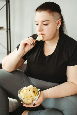 Overweight woman sitting on yoga mat eating chipsの写真素材
