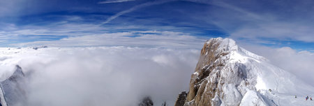 Horizontal panorama of a snow covered alpine mountain surrounded by a thick layer of clouds, beneath...