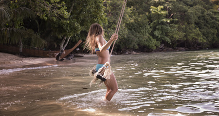 Adult Caucasian woman in blue swimsuit swinging on a rope swings by the beach.の写真素材
