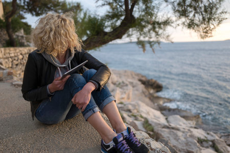 Beautiful blonde girl with curly hair is seating on the stones near of the sea with tablet in hands...