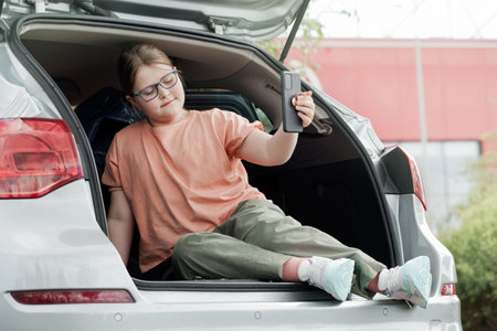 Cute preteen girl sitting in car, using smartphone ignoring her scooter on background. Kids using technology concept.の写真素材
