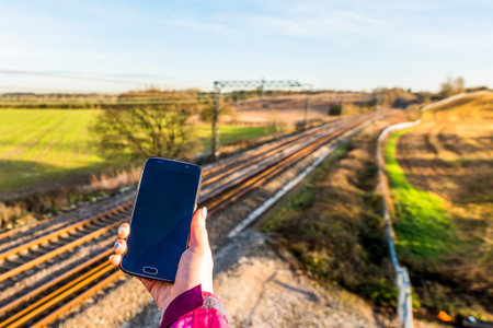 Day view of woman hand holding smartphone over UK Railroad.