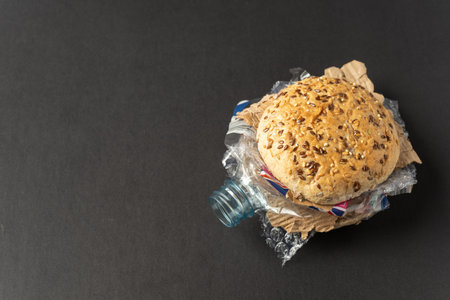 Fresh tasty burger with plastic waste and paper cardboard inside on dark background. Recycled waste...