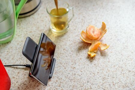 smartphone wireless charging on charging stand next to tea glass cup on kitchen tabletopの写真素材
