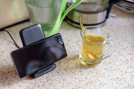 smartphone wireless charging on charging stand next to tea glass cup on kitchen tabletop