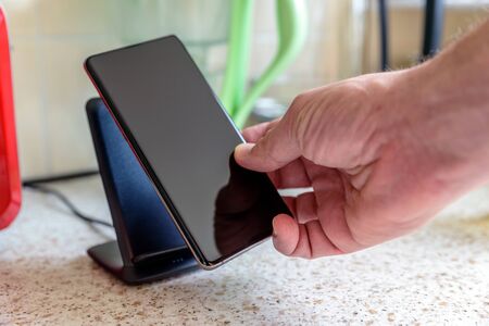 smartphone wireless charging on charging stand on kitchen tabletop. male hand placing phone to charging.の写真素材