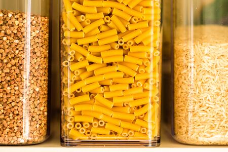 raw macaroni pasta, buckwheat and basmati rice inside clear storage container on kitchen shelfの写真素材