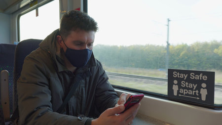 male passenger wearing face covering mask during covid-19 lockdown using phone inside train in...