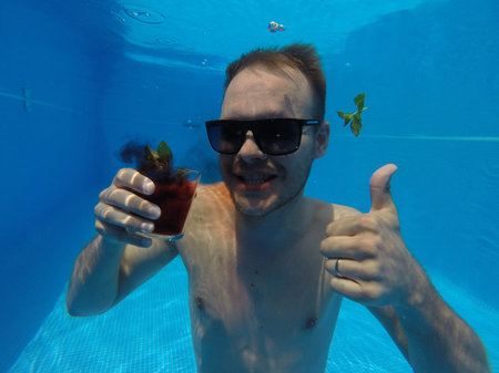 one unshaven man drinks an alcohol cocktail under the water in a pool with sunglasses in the summer,...