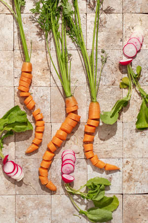 Bunch of carrot roots and ripe radishes with green leaves on stone tales of earth tones top view. Healthy eating and dieting food concept. Carrots cut in pieces remaining in natural carrot shapeの写真素材