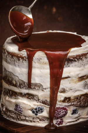 Pouring caramel sauce with spoon onto caramel cake with wild berries, cream on brown backgroundの写真素材