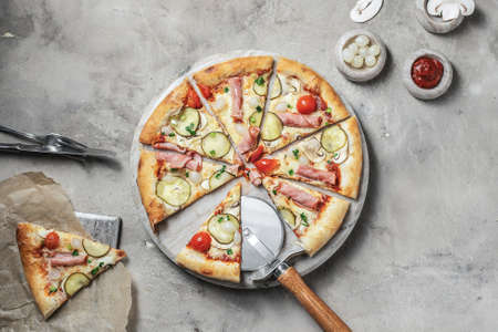 Tasty pizza with ham, cherry tomatoes, mozzarella, mushrooms, green onions and pickle cucumber on marble round tray. Garnished on grey stone backdrop with pizza ingredientsの写真素材