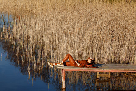 Portrait of a woman in autumn cozy orange clothes with backpack sitting on a wooden bridge near the lake with blue water and looking agains the sun. People, travel, nature concept.の写真素材