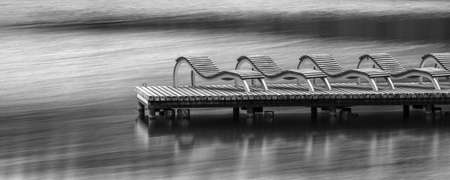 Row of empty deck chairs on dock at lake. Wooden bridge with sun-beds and observation space. Black and white photograph of relaxation, vacation and rest in nature  conceptの写真素材