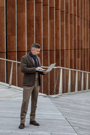 Handsome businessman using laptop on a terrace outside office, working. Grey haired man in autumn brown trendy style clothes on terrace in business area. Productive time outside the officeの写真素材