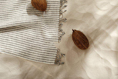 Striped linen beach towel with fringes and two coconuts on sandy beach with shadows from palm tree....
