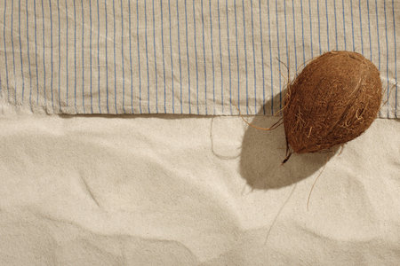 Striped linen beach towel with fringes and coconut on sandy beach with shadows from palm tree....