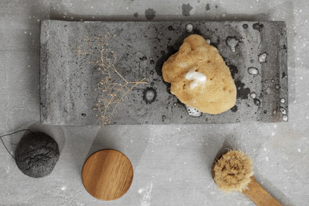 Organic sea sponge with soap foam on grey concrete tray, face creme in jar with wood lid, black charcoal konjac sponge and face brush. Bathroom accessories in contemporary bathroom interior. Daily body care, spa and wellness zero waste bathroom conceptの写真素材