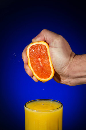 Vertical Photo of fingers squeezing fresh orange juice from a sliced orange half into a clear glass with a blue background and copy spaceの写真素材