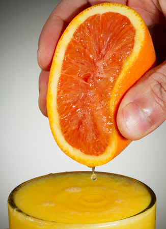 Vertical Photo of fingers squeezing fresh orange juice from a sliced orange half into a clear glass with a white backgroundの写真素材