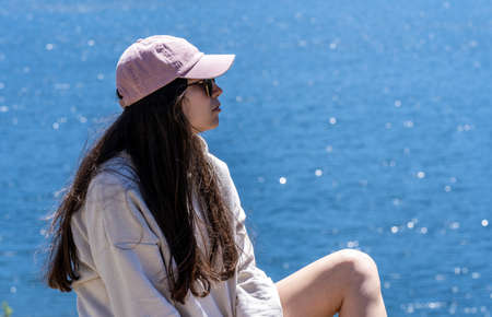 long hair brunette girl in short jeans, cap and sweatshirt enjoying the sun and the views of a mountain lake on vacation.の写真素材