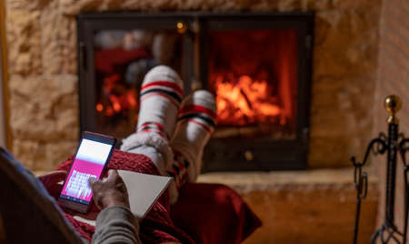 Winter night, happy woman resting by the fire with a blanket and chatting with her mobile phone, with her feet in wool socks. Cozy scene.の写真素材