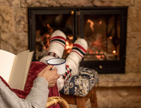 Winter night, happy woman resting by the fire with a blanket, a book and a cup of coffee, feet in wool socks Cozy scene.Winter night, happy woman resting by the fire with a blanket, a book and a cup of coffee, feet in wool socks Cozy scene.の写真素材