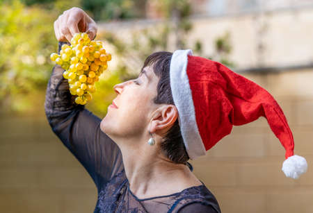 happy woman enjoys a bunch of white grapes at Christmas and wears a Santa Claus hatの写真素材