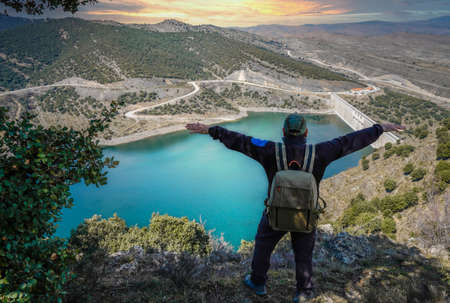 Person with backpack and open arms happily enjoying the view of a dam from the mountain.の写真素材