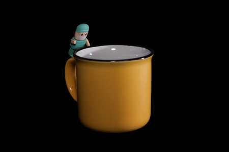 male nurse in mask and green pajamas leaning out of a yellow cup of coffee, on black backgroundの写真素材