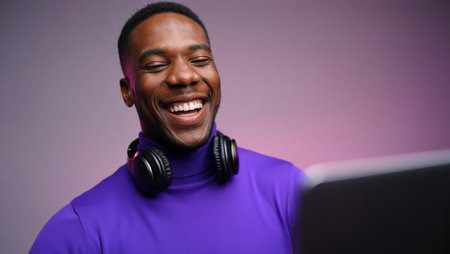 Smiling man in purple turtleneck with headphones around neck looking at laptop, enjoying online conversation or streaming.の素材