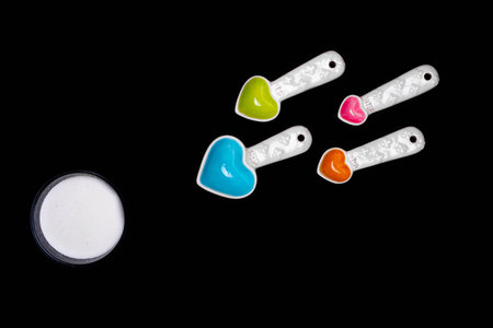 Heart shaped measuring spoons of diffferent sizes and colors facing the direction of a round bowl of sugar bringing together the ideas of love, conception and food, or sweet stuff. All on a black isolated background.の写真素材