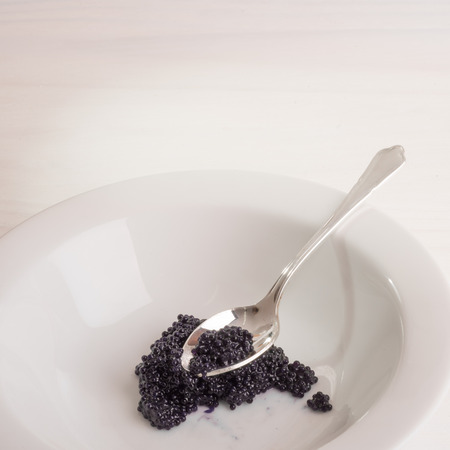 A silver spoon holds some black caviar on top of a heap of caviar inside a white bowl placed on a white wooden surface.の写真素材