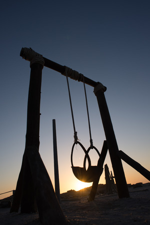 A silhouetted swing made of wooden poles, sisel rope and a tyre stand against a setting sun and dark blue sky.の写真素材