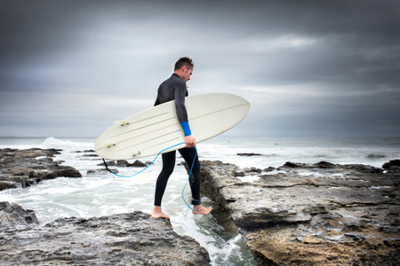 A man crosses the rocks and cravises on his way to the surf while holding a surf board in his arm.の写真素材
