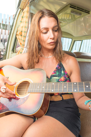 A pretty young woman sits and plays the guitar at the side door of her old classic combi van.