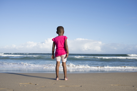 A littl girl dressed in pink stands on the beach while viewing the sea as seen from behind.の写真素材