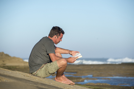 A man sits on the rocks by the ocean while reading a book and listening to music on earphones at the same time.の写真素材