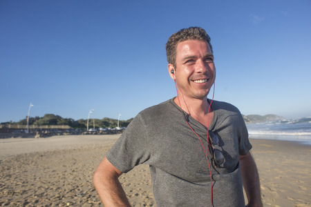 Half length portrait of a man with earphones in ears standing on the beach with a smile as he listens to music and viewing the ocean.の写真素材