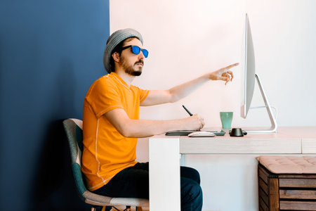 Graphic designer points screen, wears orange shirt, hat and sunglasses, blue background.