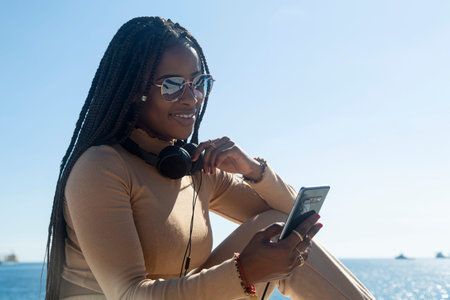 Young black afro woman smiling looking at her cellphone, outdoors with blue sky and sea background....