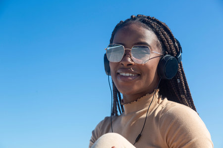 Closeup portrait of young black afro woman smiling with headphones and blue sky background, pretty...